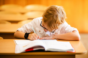 Seven years old child with glasses writing his homework at school. Boy studing at table on class background