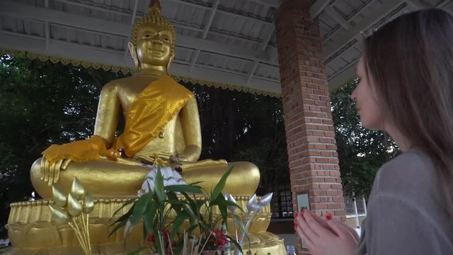 Beautiful, young woman with long blond hair in front of the altar and a golden Buddha statue. Buddhist temple in the open space.