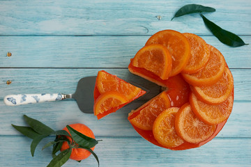 Top view and close-up of a cold homemade orange pie with candied orange on the surface and orange jam accompanied by a piece of orange pie and an fresh orange with green leaves ona blue wooden backgro