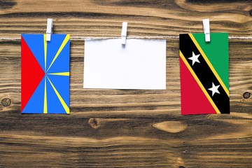 Hanging flags of Reunion and Saint Kitts And Nevis attached to rope with clothes pins with copy space on white note paper on wooden background.Diplomatic relations between countries.