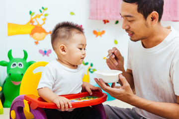 Obraz premium Father acting Mom feeding his son baby 1 year old on chair