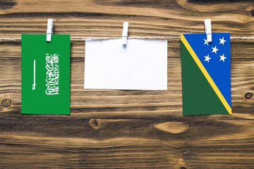 Hanging flags of Saudi Arabia and Solomon Islands attached to rope with clothes pins with copy space on white note paper on wooden background.Diplomatic relations between countries.