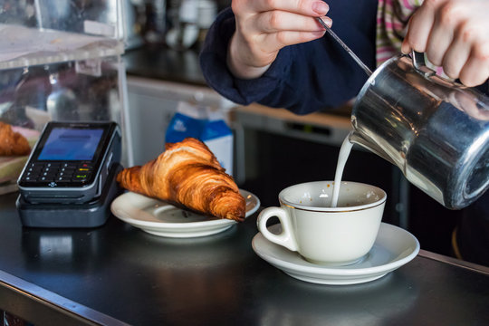 Barista female hands pouring hot milk on a cup to make a cappuccino with a croissant and a contactless post in the background.