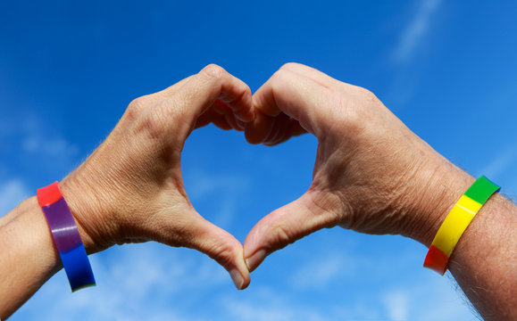 Man and womans hands forming a heart wearing rainbow wristband colors against a blue sky