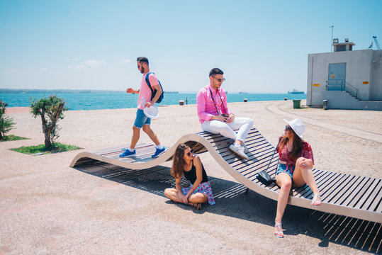 Four tourist friends having fun and taking pictures on a modern abstract bench paced on a seaside marine port on a sunny day. Enjoying the beautiful blue and turquoise sea.