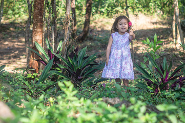 Pretty three year old girl, white violet dress, red flower in her hair, smiles and shows a little flower in her hand, is