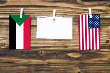 Hanging flags of Sudan and United States attached to rope with clothes pins with copy space on white note paper on wooden background.Diplomatic relations between countries.