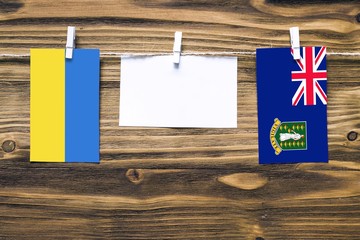 Hanging flags of Ukraine and British Virgin Islands attached to rope with clothes pins with copy space on white note paper on wooden background.Diplomatic relations between countries.