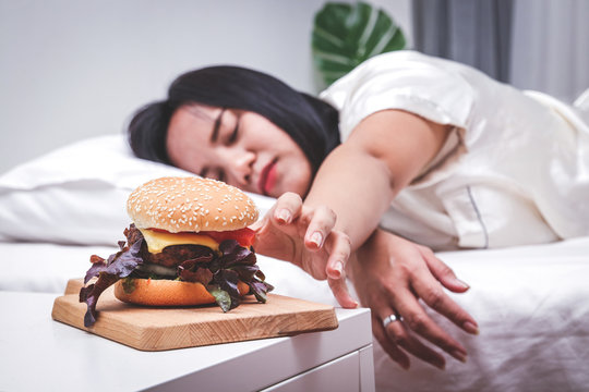 Fat woman lying on the bed Reaching for an empty hamburger on the table to eat on the bed. Concept of weight loss