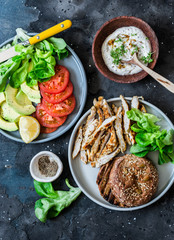 Healthy diet lunch - grilled chicken fillet, rye seed bun, fresh tomatoes, lettuce, avocado and yogurt mustard sauce on dark background, top view