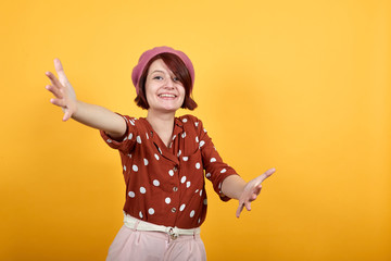 Caucasian attractive young brunette woman over isolated yelllow background on funny pink hat looking at camera smiling with open arms for hug. Cheerful expression embracing happiness.