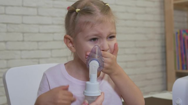 A little girl takes a breath in an inhalation mask. The girl resists and does not want to do the procedure.