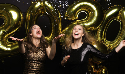 Two happy girls in shiny dresses posing while standing with gold colored 2020 number balloons on black background.