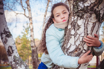 Cute girl with long hair in blue down jacket hugging birch with her hands on an autumn day.
