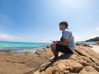 Asia lesbian of  woman 20-30s in hair cut using and hand holding smartphone connected to digital camera selfie photo take yourself an sitting on a rock at the beach