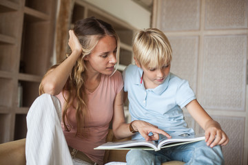 young beautiful mother and teenage son are sitting on an armchair and reading a book. blond family in home interior