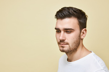 Fototapeta premium Studio shot of attractive young European man with bristle looking down with shy smile posing in white t-shirt against blank wall background with copy space for your advertising information