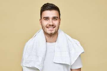 Studio image of attractive cheerful young European male with stubble and white towel around his neck smiling broadly at camera, going to shave his face in bathroom in the morning before work - Powered by Adobe