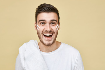 Obraz premium Studio shot of emotional excited young bearded guy posing isolated in t-shirt and towel on his shoulder going to shave and take shower after training in gym, stripes of white foam on his cheeks