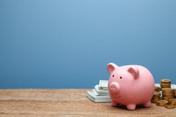 Pink piggy bank and money with banknotes and coins on a wooden table and blue background. Copy...