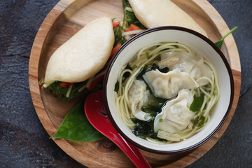 Round bamboo serving tray with a bowl of wonton noodle soup and baozi, view from above, close-up