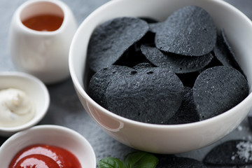 Close-up of black potato chips served in a white bowl with dips, selective focus