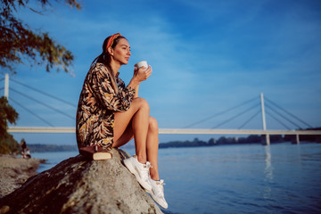 Side view of charming brunette in floral dress and with headband sitting on rock on shore and drinking beverage from mug. In background is bridge.