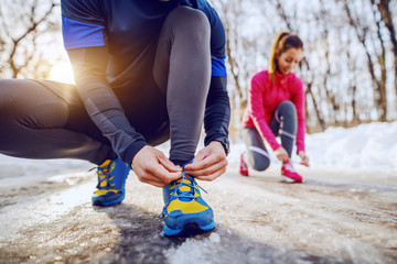 Fit caucasian man in sportswear crouching on country road and tying shoelace. In background is his female friend tying shoelace, too. Wintertime. Outdoor fitness concept.