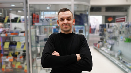 Fototapeta na wymiar Portrait of best a Professional Expert Consultant Smiles and Looks into Camera as Stands in the Bright, Modern Electronics Store Full of Latest Models of TV Sets, Cameras, Table 