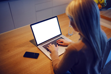 Rear view of attractive blonde woman typing on laptop while sitting at dining table at home. On table are smart phone and glass of water.