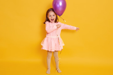 Horizontal shot of little girl with purple balloon isolated over yellow studio background, female child shouting somethig, celebrating herbirtday, kid wearing rosy dress and having dark hair.