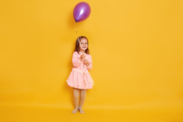 Studio shot of little girl wearing rose dress playing with purple balloon isolated over yellow background, having birthday, having fun with balloon, cute female child with dark hair. Childhood concept