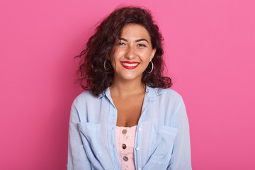 Horizontal shot of beautiful woman, brunette girl stands smiling against rosy backgeound, having dark wavy hair and clean skin, female wearing stylish blue shirt, looking directly at camera. © sementsova321