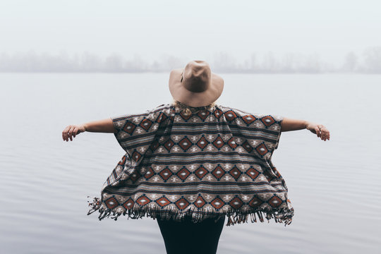Woman in hat and poncho standing on the river bank and looking towards autumn forest covered with morning fog