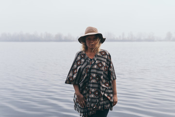 Woman in hat and poncho standing on the river bank and looking towards autumn forest covered with morning fog