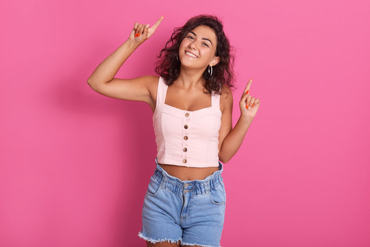 Portrait of adult happy brunette woman pointing up with her index fingers, standing isolated over pink background, expressing happyness and joy, female with darkwavy hair, wearing stylish cloth.