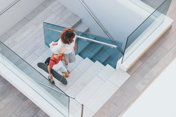 Trendy mother and son walking on stairs in a modern city context. Young boy with skateboard goes up the ladder with mum. Top view of people walks in a modern mall. Family scene at the shopping center - Powered by Adobe