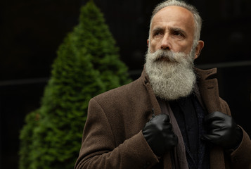 Handsome confident mature man with beard stands in coat on street with copy space for your text message or advertising, fashionable rich male dressed in expensive clothes posing outdoors.