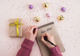 New year Goals Concept 2020. A woman writes plans in a notebook on a table with a festive decor. Stylish composition in light pink and gold colors. Top view, flat lay
