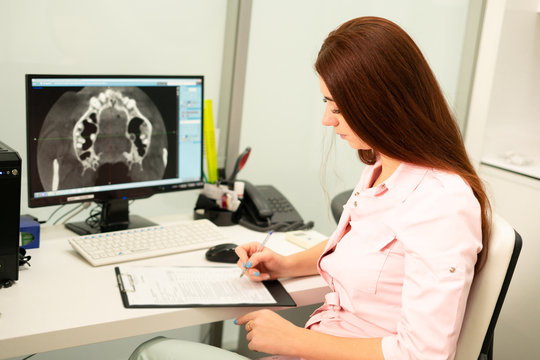 A female dentist doctor is sitting at a table, on a computer a CT scan of the jaw. The doctor is dressed in professional clothes.
