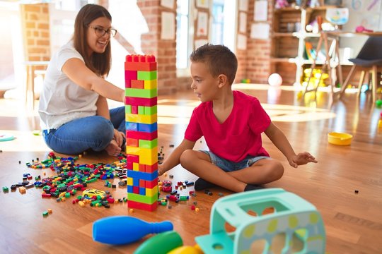 Young beautiful teacher and toddler playing with building blocks around lots of toys at kindergarten