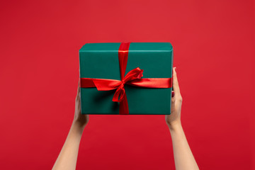 cropped view of woman holding green wrapped Christmas present with ribbon isolated on red background. Christmas concept
