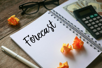 Top view of calculator,dollar bank notes,glasses, trash paper,pen and notebook written with Forecast on wooden background.