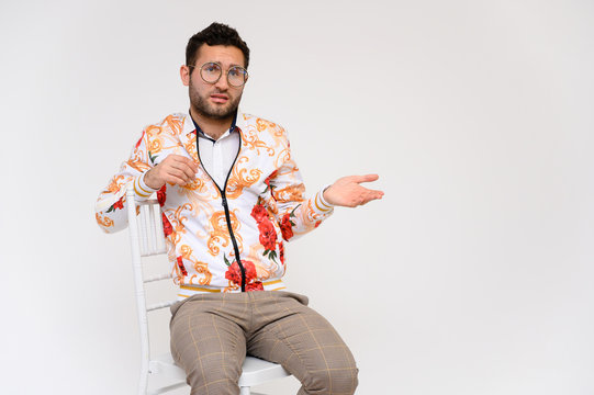 Men's fashion concept. Portrait of a handsome male model, showing hands, wearing a white jacket with a floral pattern, posing on a white background, sidmint on a chair. Black hair. Close Studio Shot