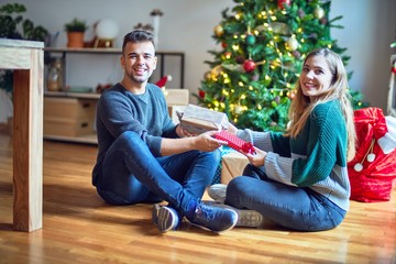 Young beautiful couple smiling happy and confident. Man surprise woman with gift around christmas tree at home