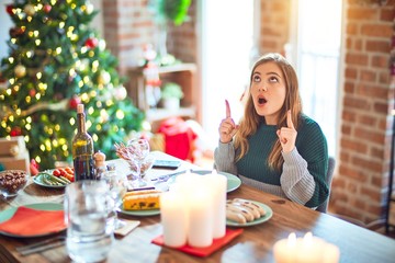 Young beautiful woman sitting eating food around christmas tree at home amazed and surprised looking up and pointing with fingers and raised arms.
