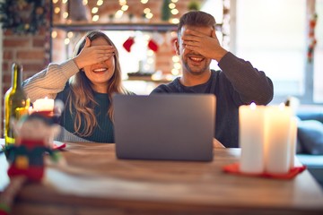 Young beautiful couple sitting using laptop around christmas decoration at home smiling and laughing with hand on face covering eyes for surprise. Blind concept.
