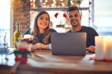 Young beautiful couple sitting using laptop around christmas decoration at home winking looking at...