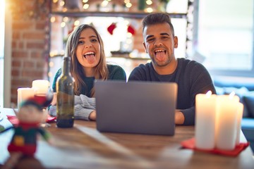 Fototapeta premium Young beautiful couple sitting using laptop around christmas decoration at home sticking tongue out happy with funny expression. Emotion concept.