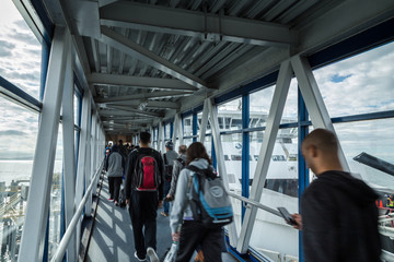 A passenger ferry terminal gangway with travellers walking with their luggage towards a ferry waiting to depart. - obrazy, fototapety, plakaty